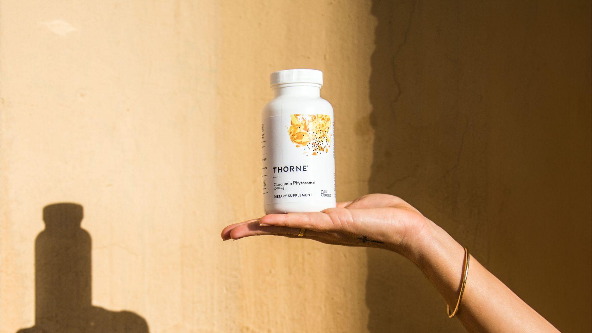 A hand holding a bottle of Thorne Curcumin Phytosome against a sunlit wall